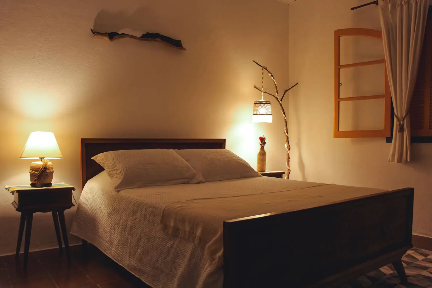 Quarto aconchegante com cama de casal, abajur aceso e janela de madeira