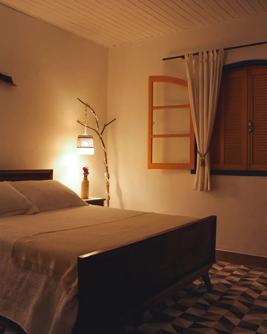 Cama de casal e luminária de chão junto à janela de venezianas