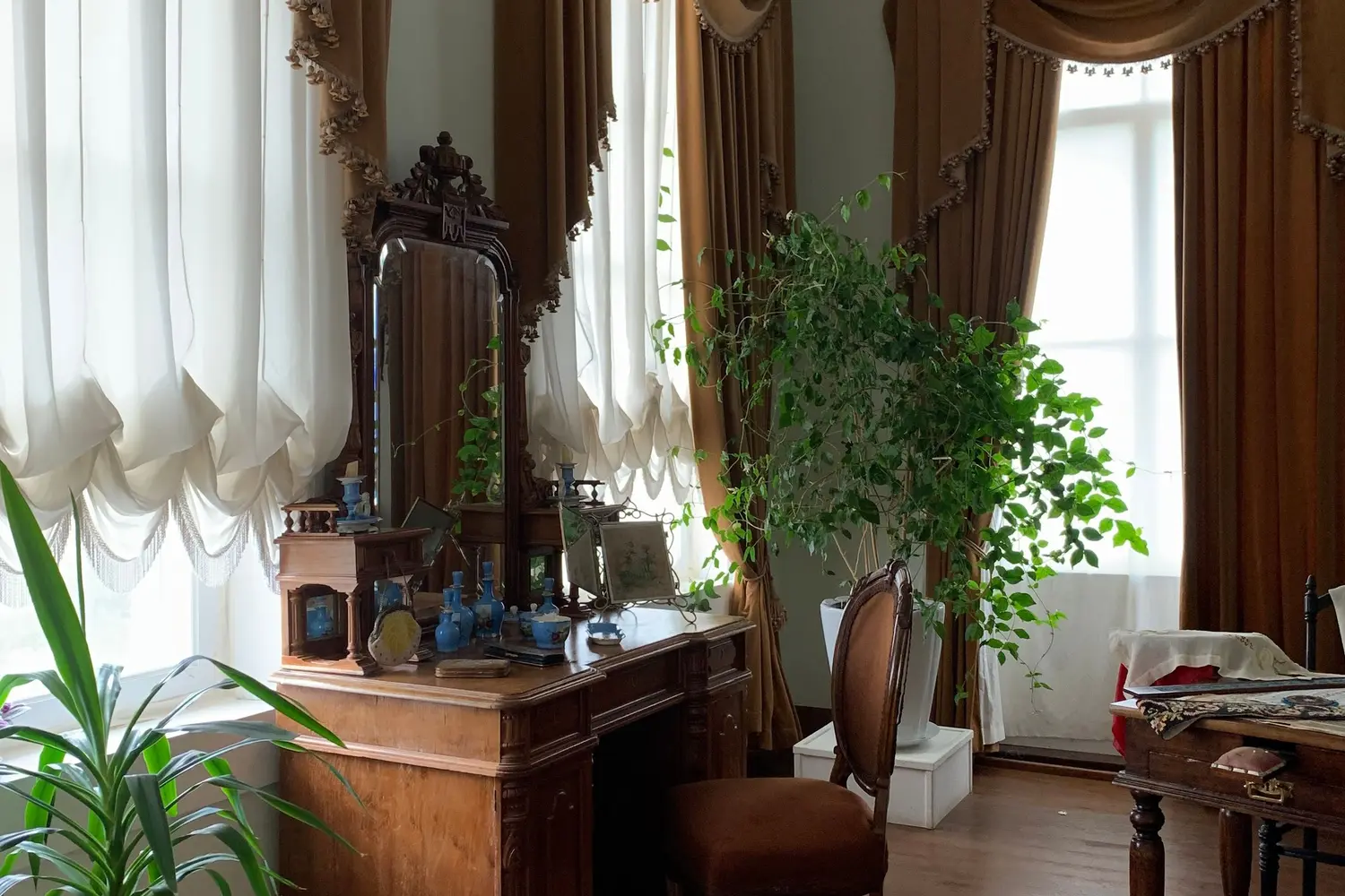 Escrivaninha antiga com espelho, cadeira e planta, junto a janelas altas com cortinas
