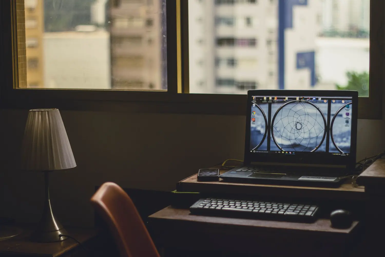 Mesa de trabalho com notebook, teclado e abajur junto à janela